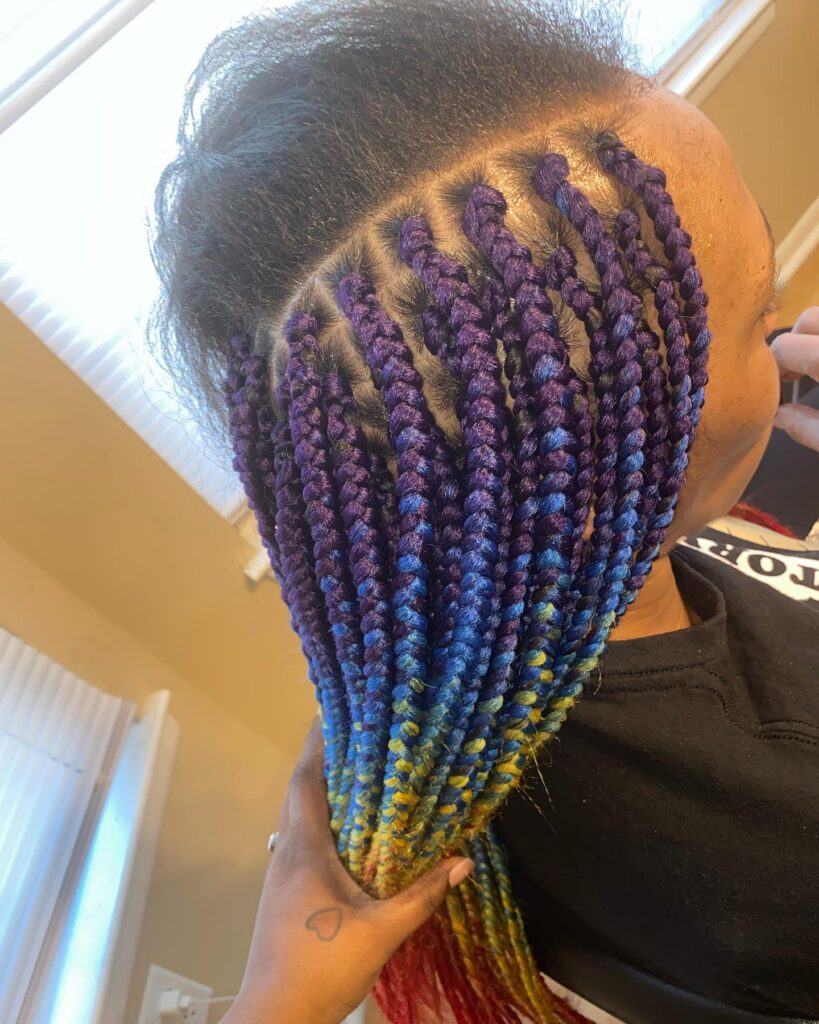 A woman showing off long box braids that ombre from purple to blue and yellow with neat square partings.