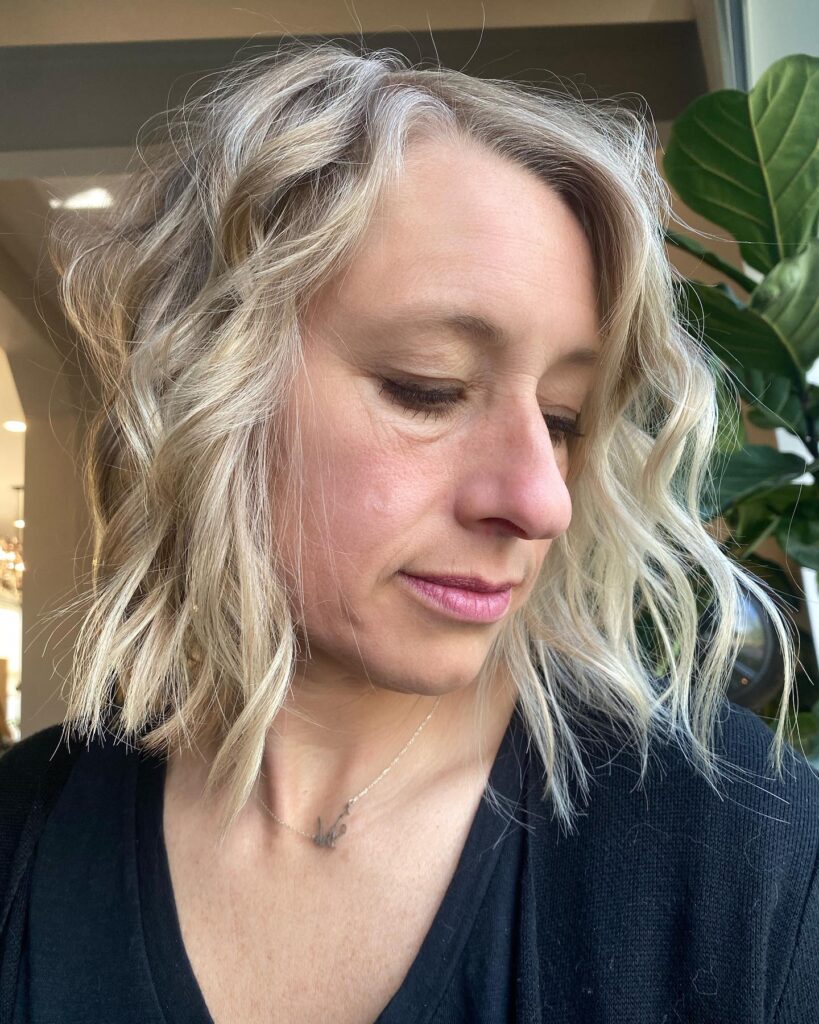 A woman with a wavy shoulder length lob featuring a mix of sandy blonde and silver highlights with textured layers.