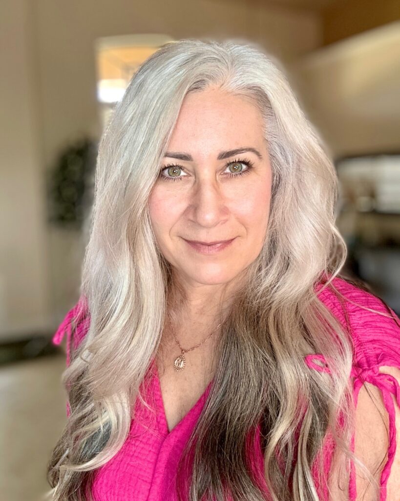 A woman with long wavy silver hair and dark grey lowlights wearing a pink shirt.