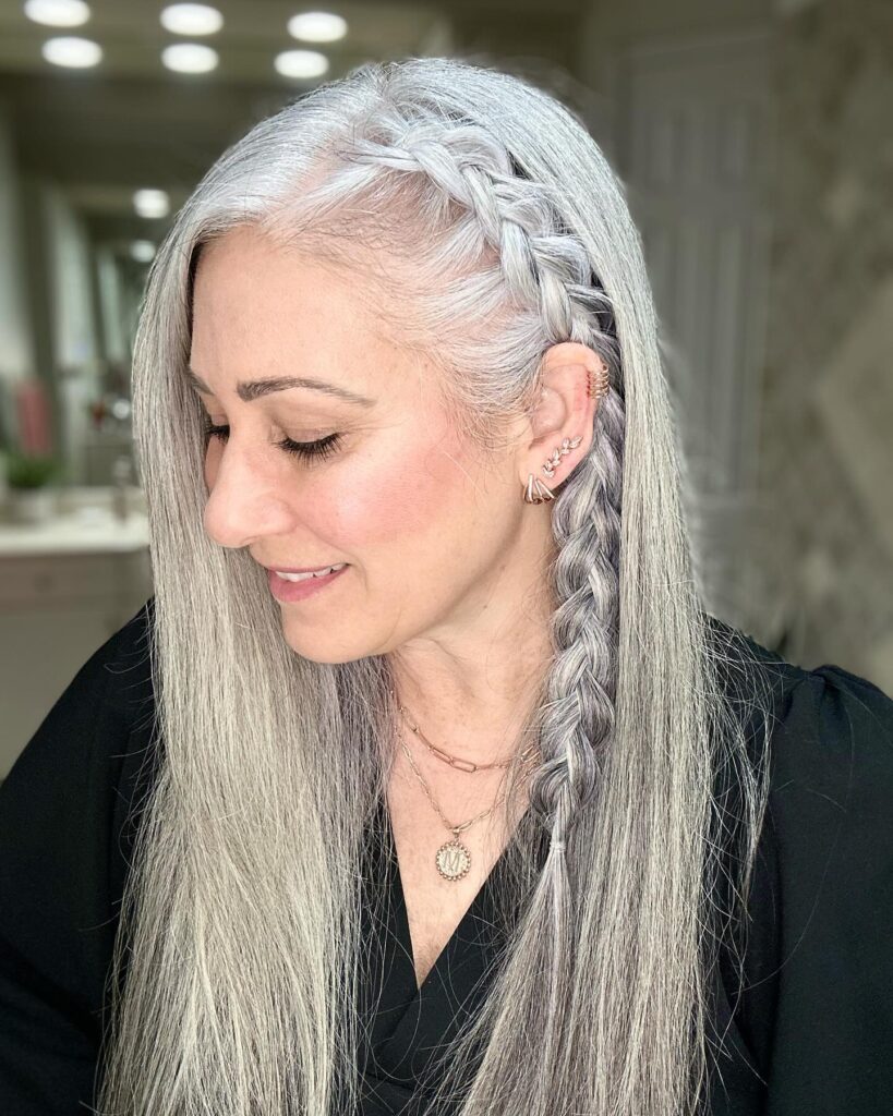 A woman with long straight silver hair featuring a thick side braid and elegant gold jewelry.