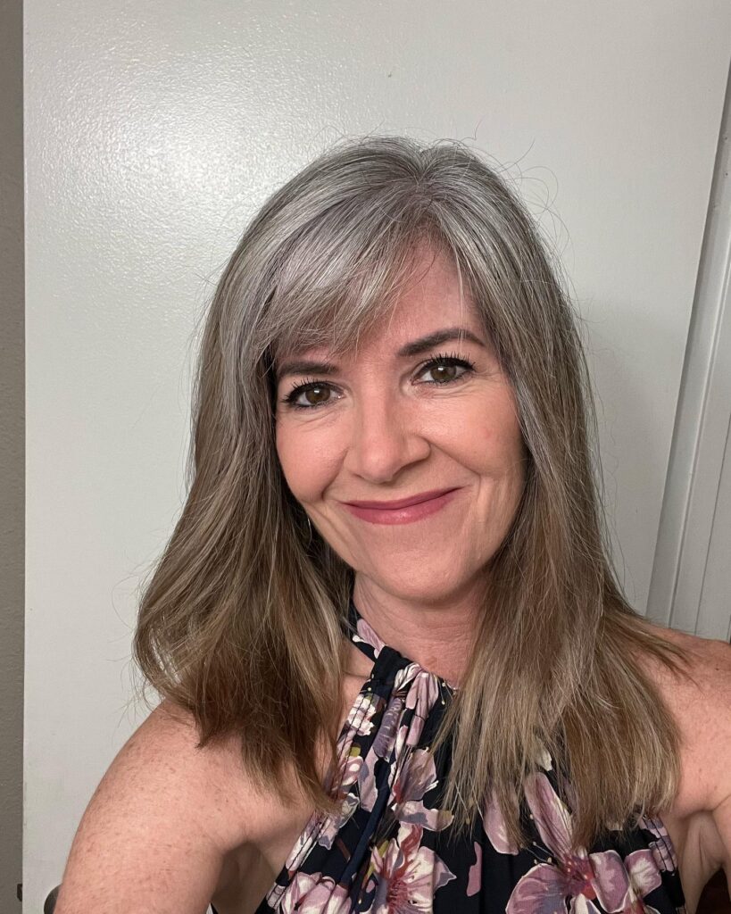 A woman with straight mid length bright silver hair and a wispy fringe smiling for a selfie.