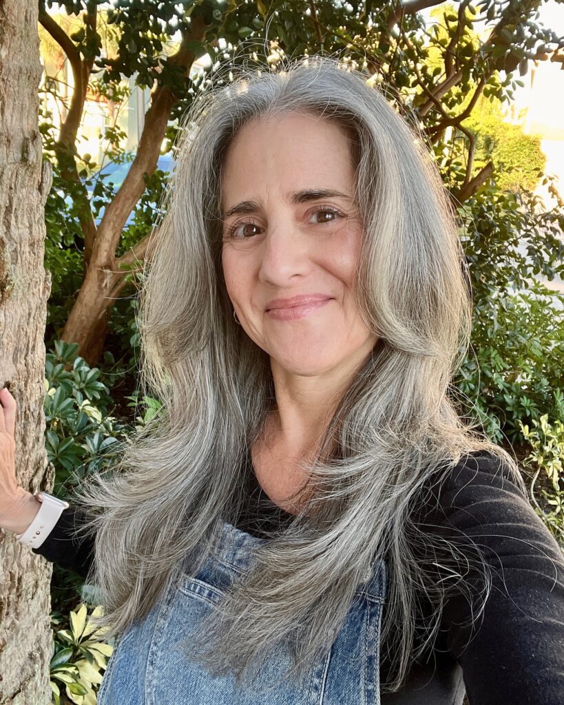 A woman with long layered silver hair and face framing pieces smiling outdoors next to a tree.