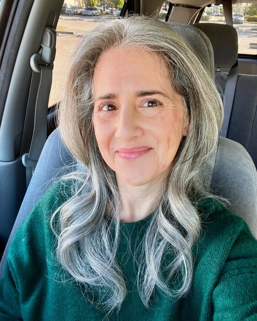 A woman with long wavy salt and pepper hair smiling while sitting in a car wearing a green sweater.