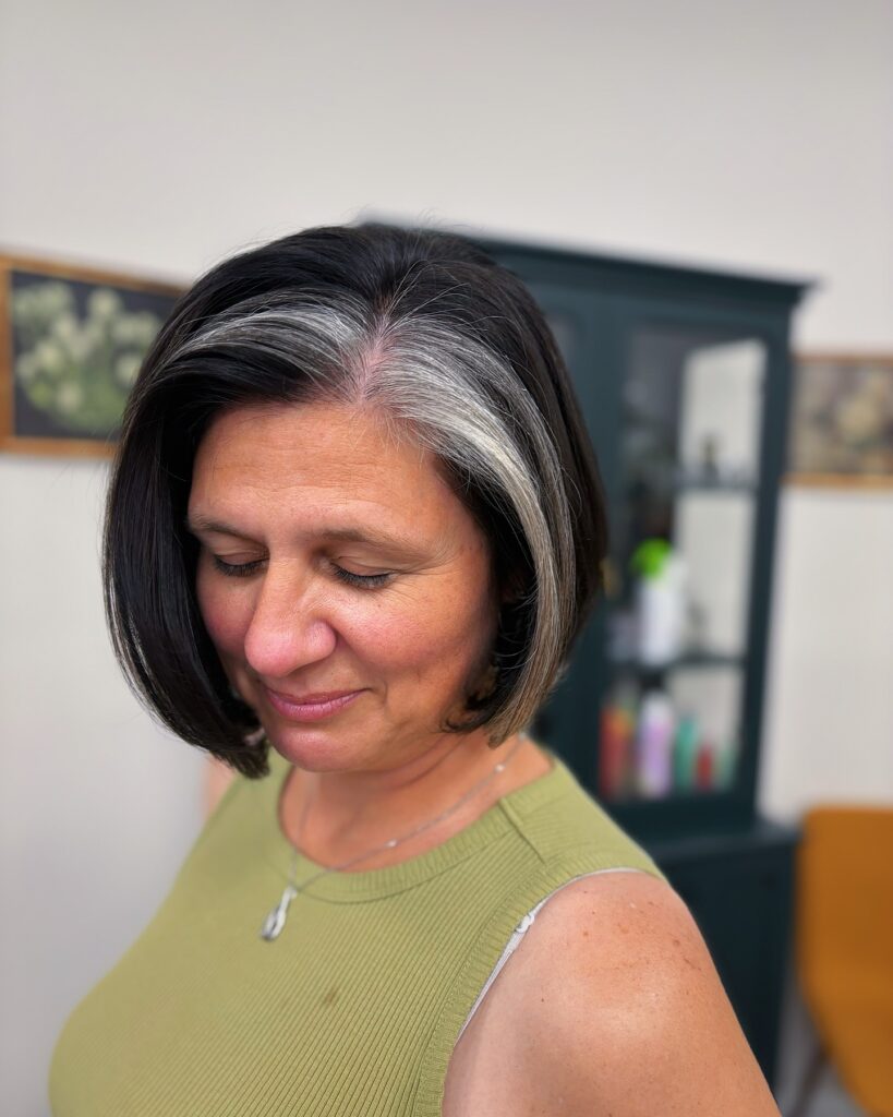A woman with a dark brown chin length bob featuring a prominent silver streak at the front hairline.