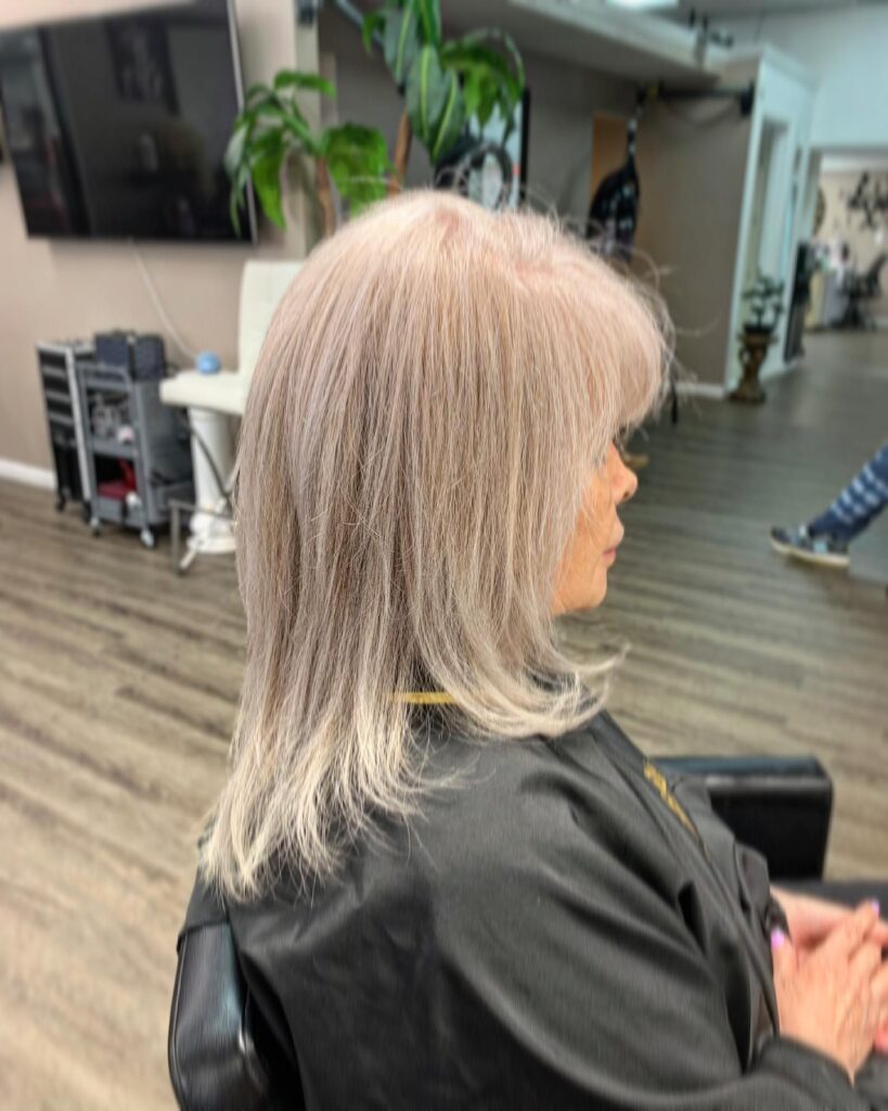 A side view of a woman with mid length champagne silver hair featuring wispy bangs and slightly flipped ends.