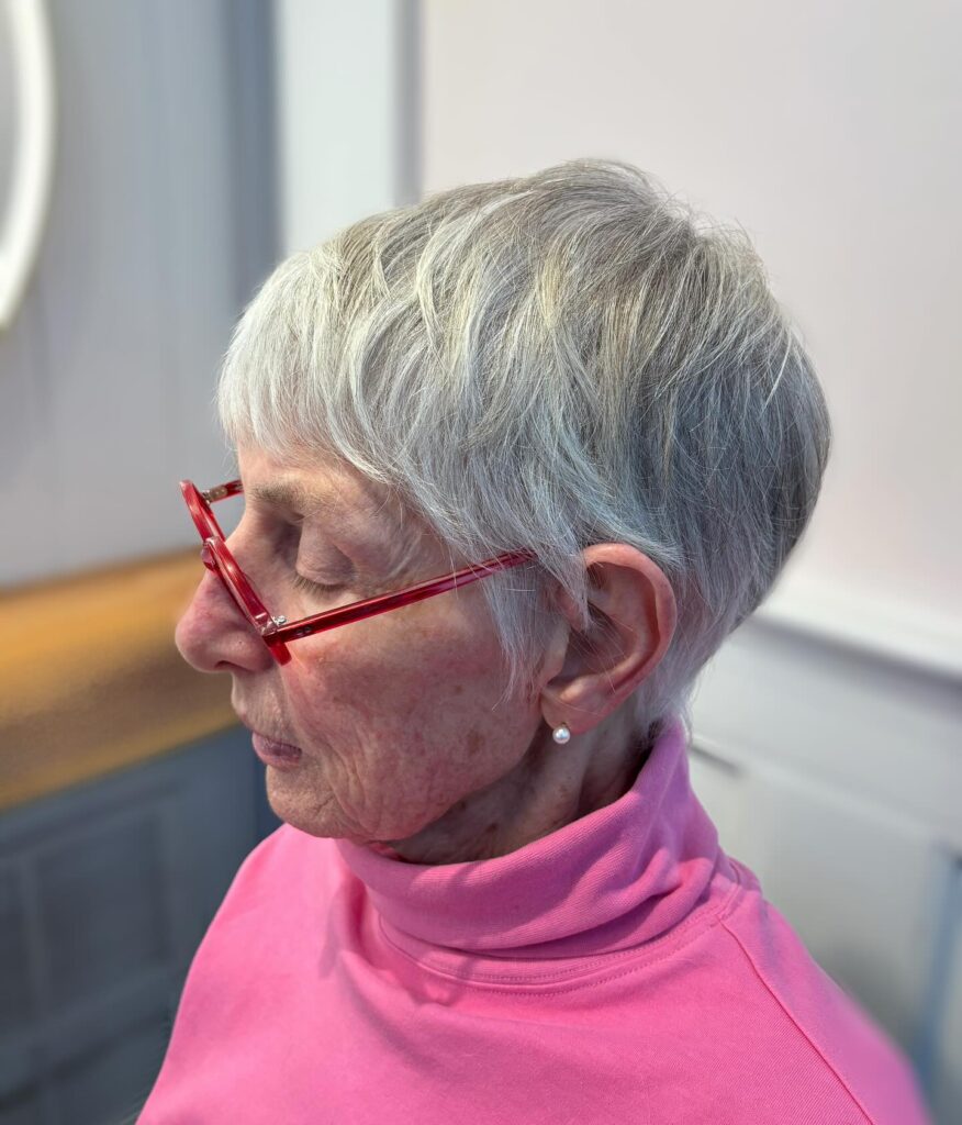 A woman with a very short silver pixie cut and micro bangs wearing red glasses and a pink turtleneck.