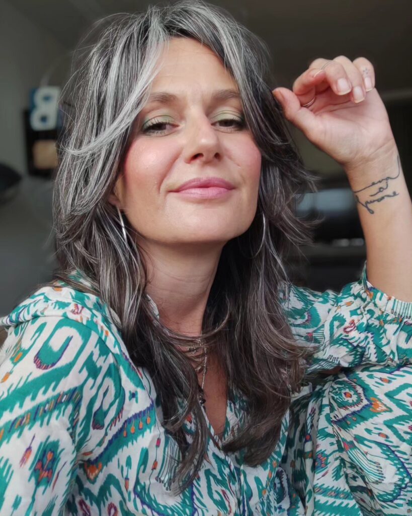 A woman with a layered gray shag hairstyle featuring bright silver face framing highlights and wavy texture.