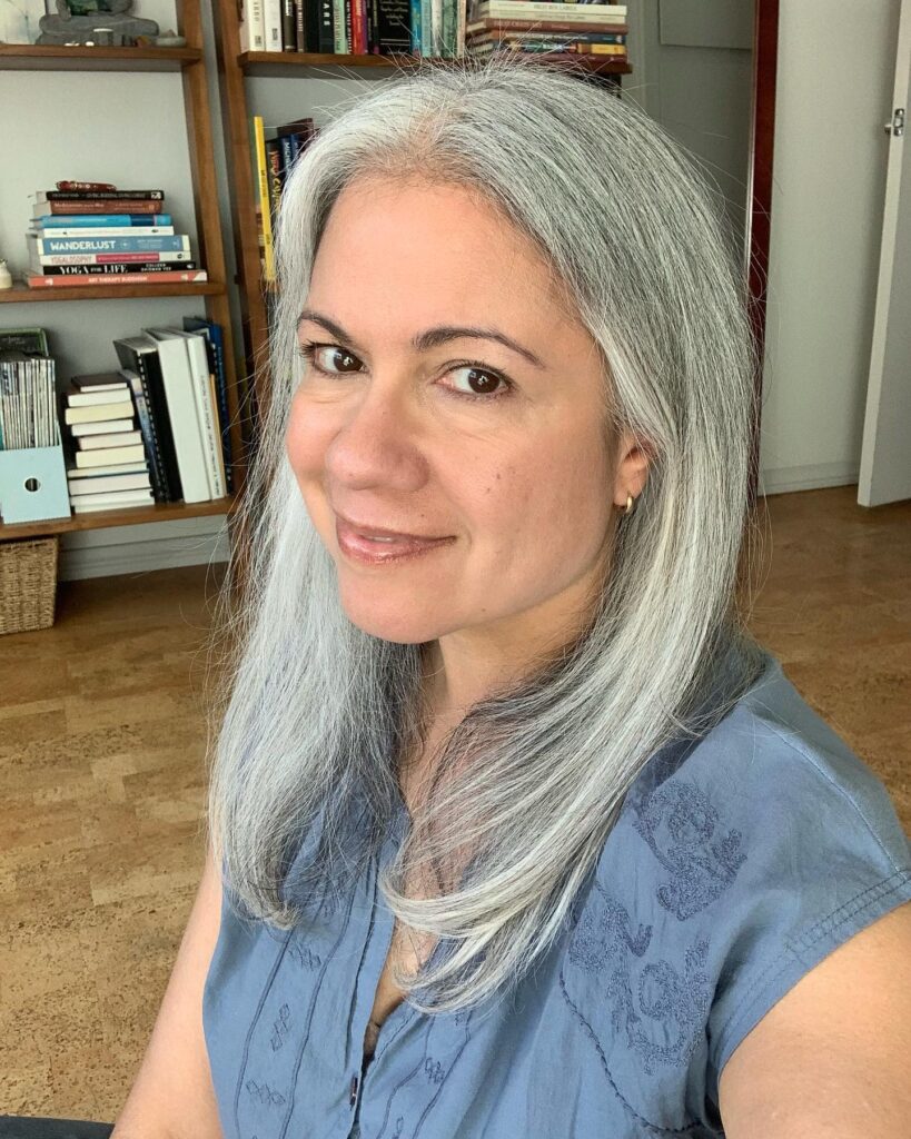 A woman with mid length straight silver hair and long layers wearing a blue sleeveless shirt.