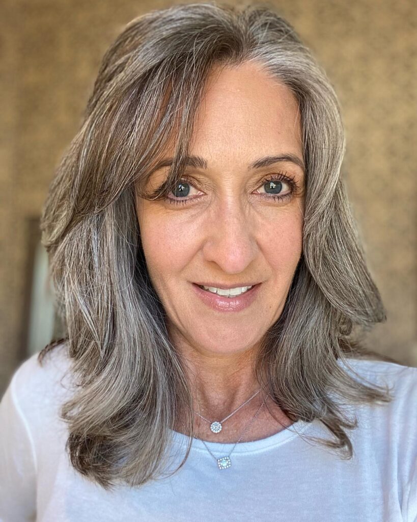 A woman with shoulder length layered silver and gray hair and side swept bangs smiling for a close up photo.
