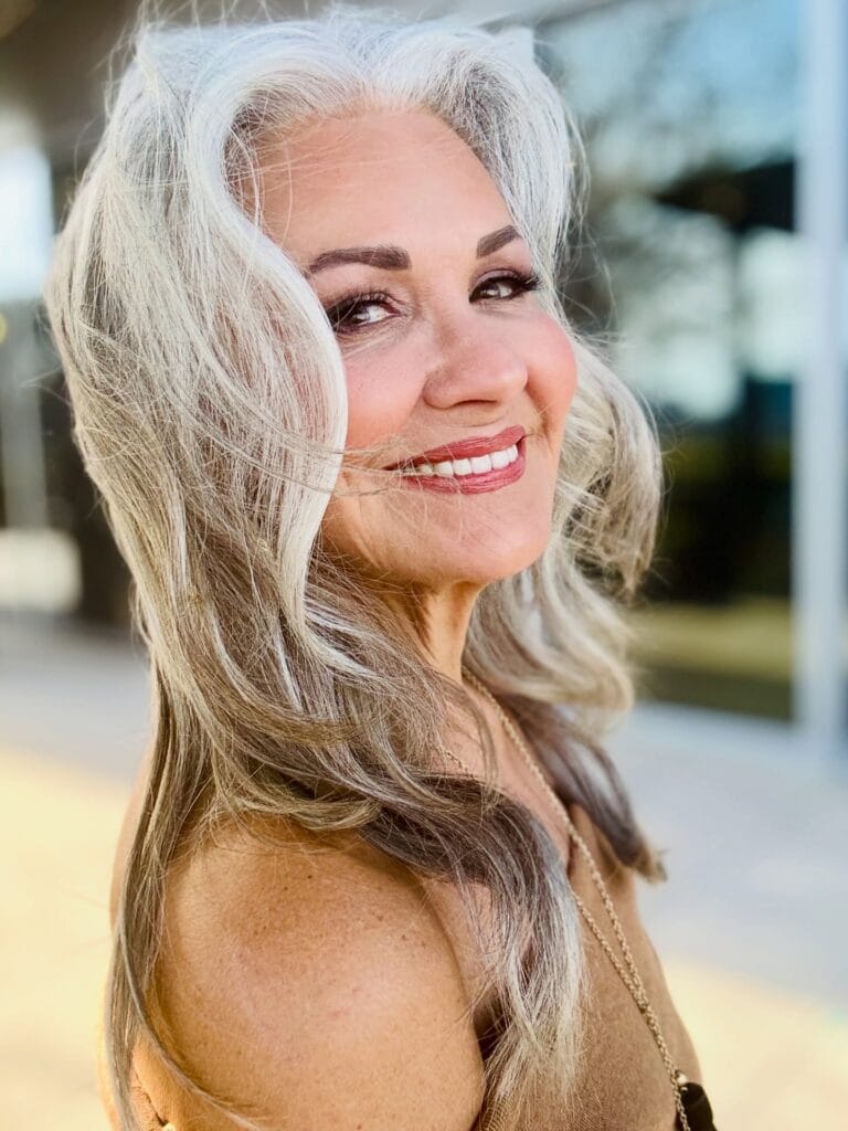 A woman smiling with long wavy silver and blonde hair featuring voluminous face framing layers.