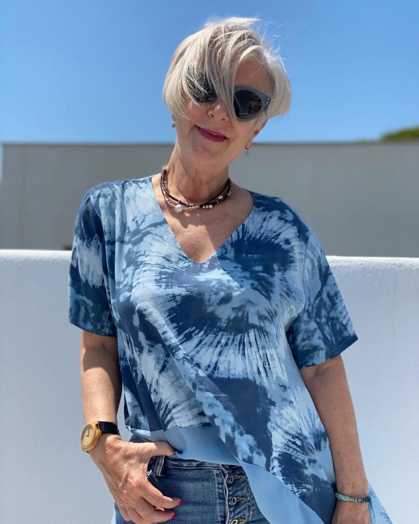 A woman with a short silver asymmetrical pixie cut and long side bangs wearing dark sunglasses and a blue tie dye shirt.