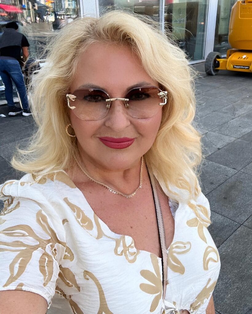 A woman with mid length layered blonde hair and soft waves wearing rimless sunglasses and a patterned white top.