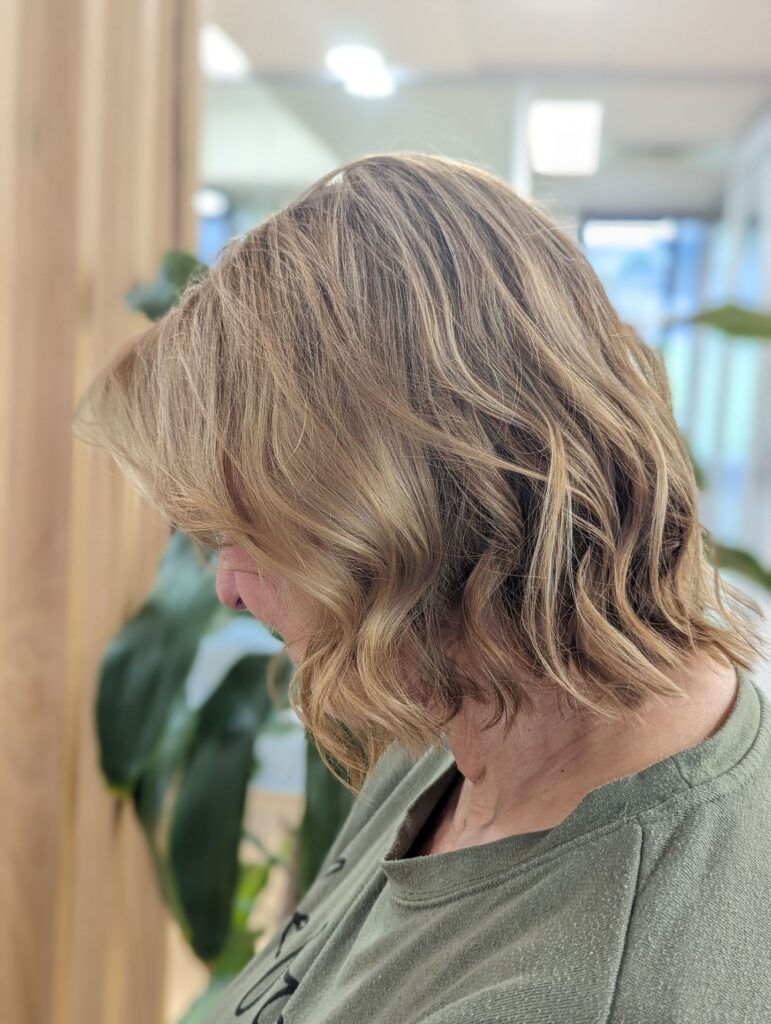 A woman showcases a neck length bob haircut with soft waves and multi dimensional honey blonde highlights.