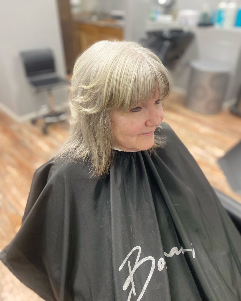 A woman with a medium length ash blonde shaggy haircut featuring full bangs and textured layers.