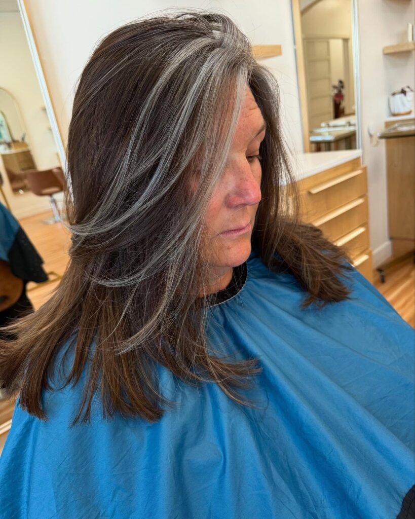 A woman with long brunette hair featuring natural silver highlights and soft face framing layers.