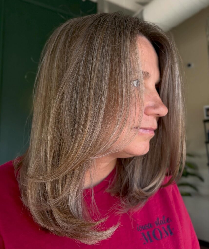 A woman showcases a straight mid length hairstyle with honey blonde highlights and soft layers at the ends.