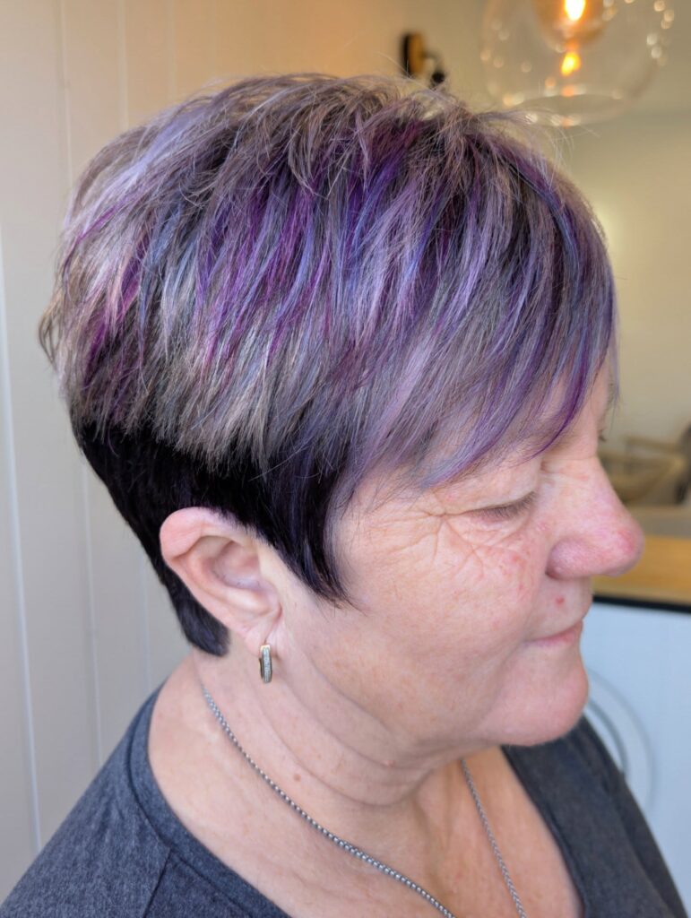 A woman with a short textured pixie cut featuring a mix of vibrant purple and silver highlights over a dark base color.
