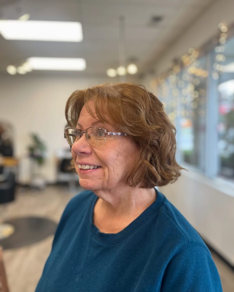 A smiling woman with glasses wearing a wavy caramel brown layered bob with soft bangs in a bright salon setting.