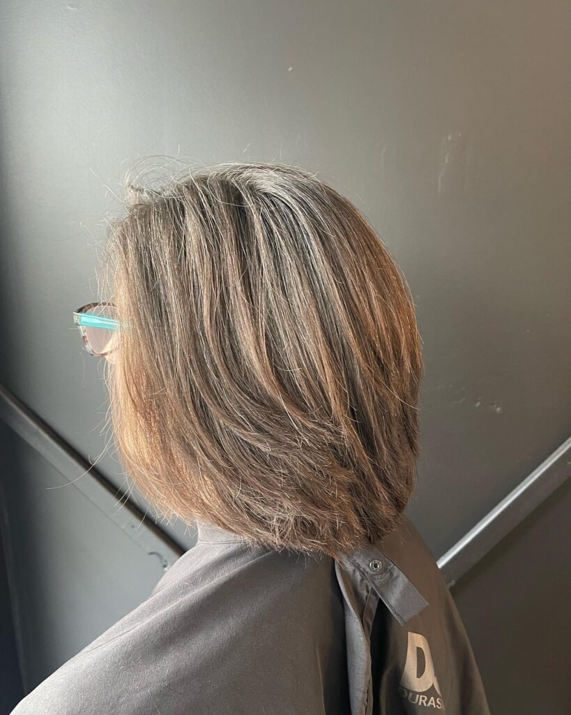 A woman viewed from behind, wearing glasses and a salon cape, showcasing a shoulder-length layered dark brown bob with natural gray highlights.