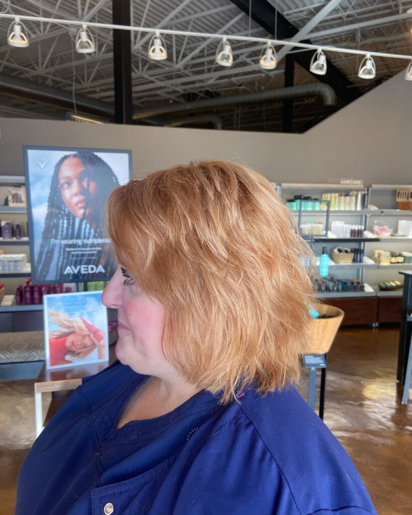 A side profile view of an older woman with a textured copper short layered bob haircut styled with movement and volume in a hair salon setting.