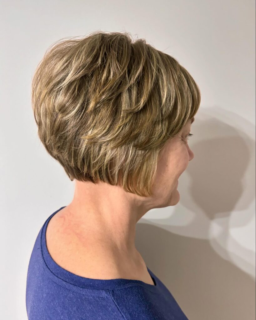 A close-up side view of a layered stacked inverted bob haircut with brown and blonde highlights on an older woman with a slight wave in her hair.