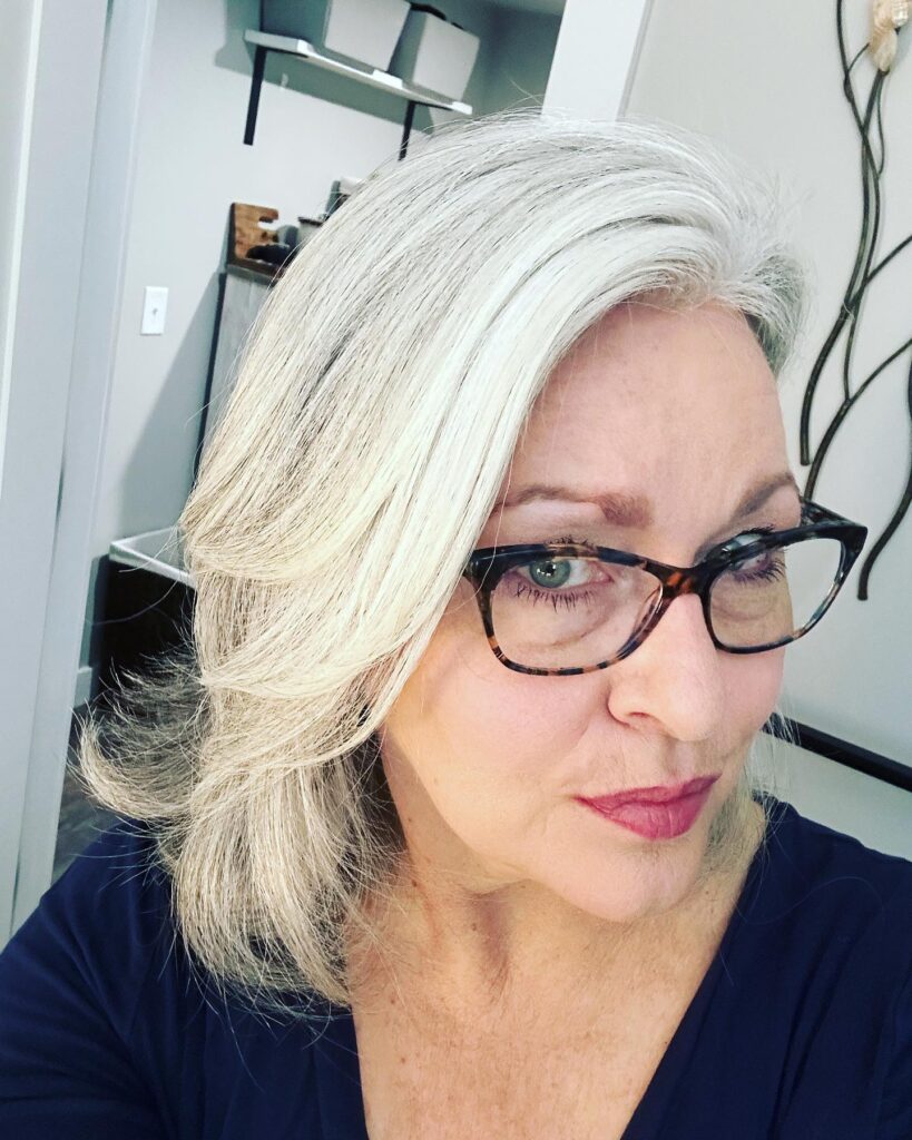 A woman with stylish glasses wearing a luminous silver shoulder length lob with swept back face framing layers.