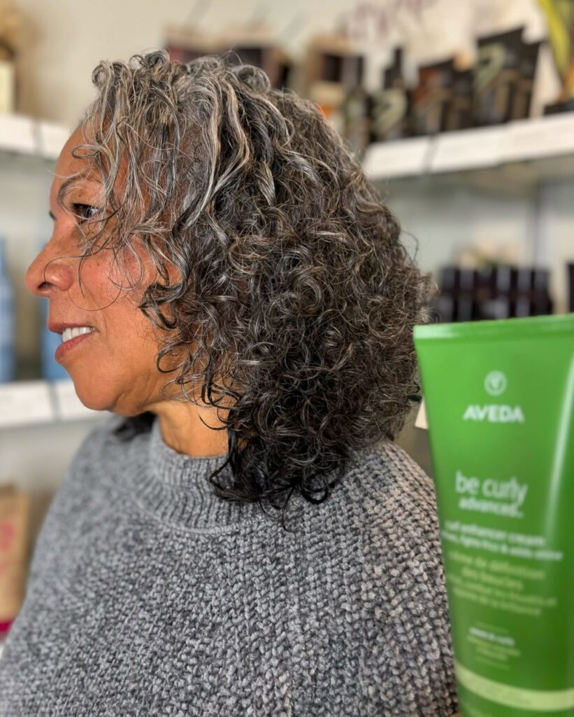 A side profile view of a woman with a natural salt and pepper curly bob haircut.