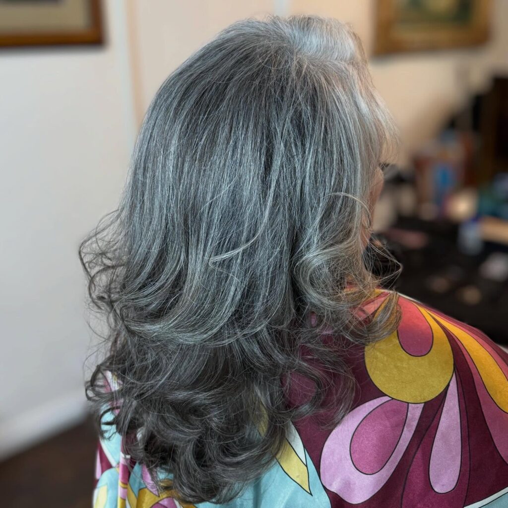 A back view of a woman with mid length layered silver hair featuring soft curls and charcoal lowlights.