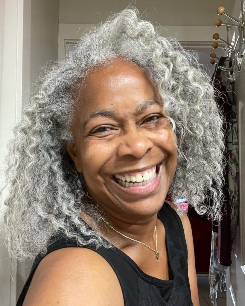 A woman with a big smile wearing a shoulder length natural silver hairstyle with soft waves and curls.