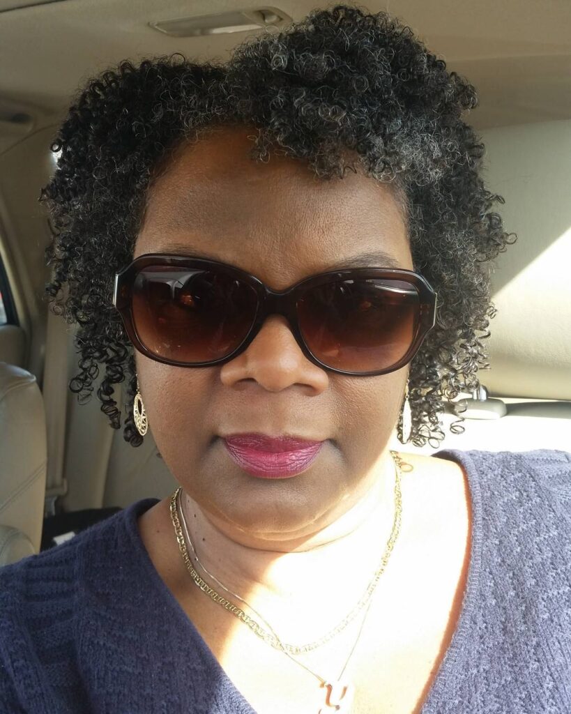 A woman wearing sunglasses with an ear length salt and pepper hairstyle featuring tight natural coils.