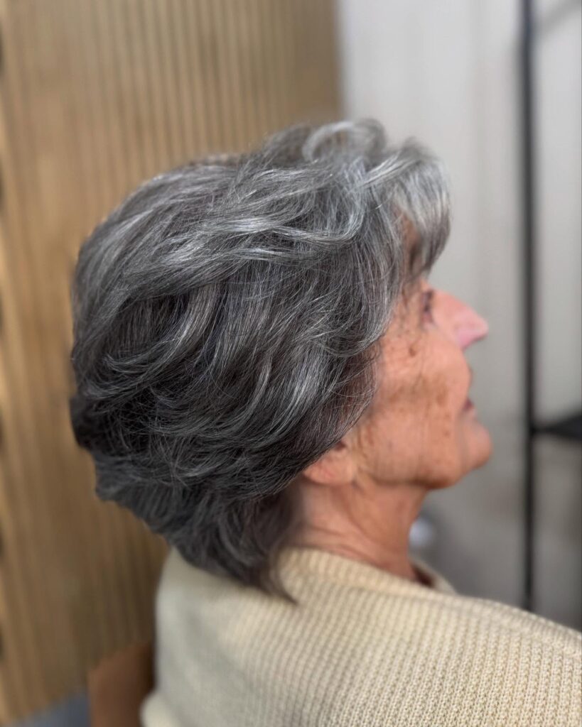 A side profile of a woman showing a feathered charcoal grey hairstyle with swept back layers and a tapered neckline.