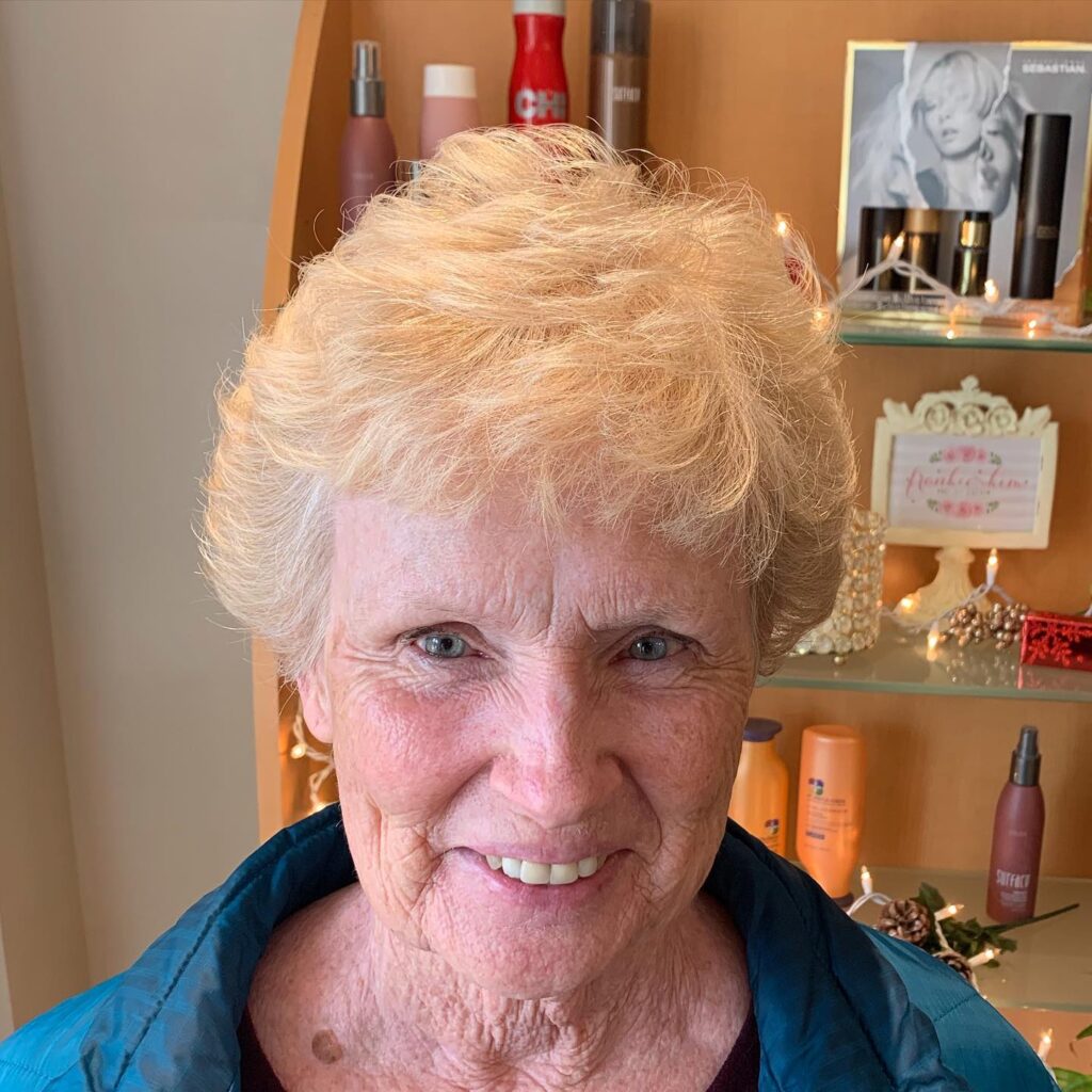 A smiling woman with an oval face shape shows off a short voluminous strawberry blonde hairstyle with feathered layers and wispy bangs.
