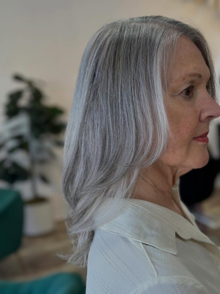 A woman with natural gray shoulder-length hair and subtle face framing layers is in profile wearing a cream blouse.