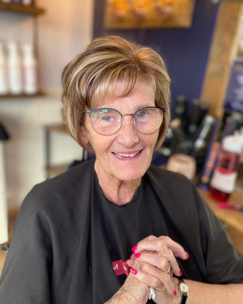 An older woman with a rounded golden brown bob featuring soft blonde highlights and light wispy bangs.