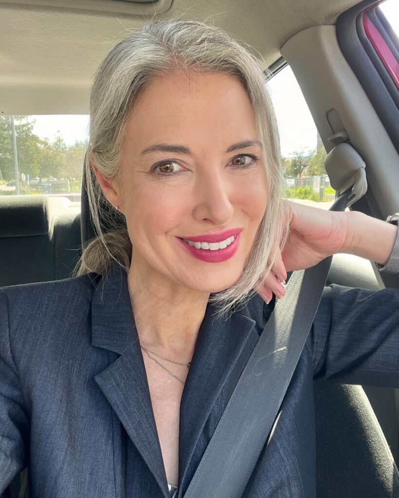 A woman with long straight silver hair styled in a low ponytail with loose strands framing her face.