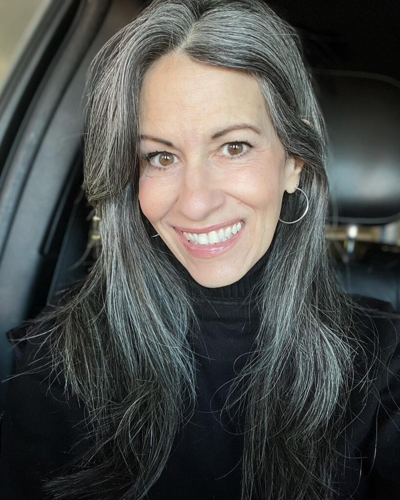 A woman with long, layered salt and pepper hair in a naturally straight style smiling widely.