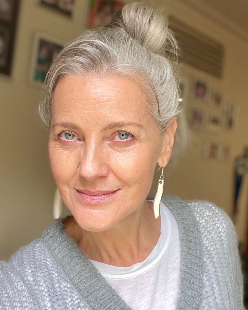 Close-up portrait of a mature woman with silver hair in a bun, wearing a gray sweater and white earrings, softly smiling indoors.