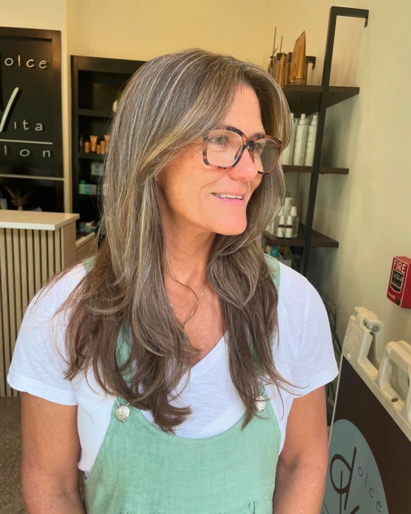 A woman with long light brown hair featuring silver highlights and face framing layers wearing tortoise shell glasses.