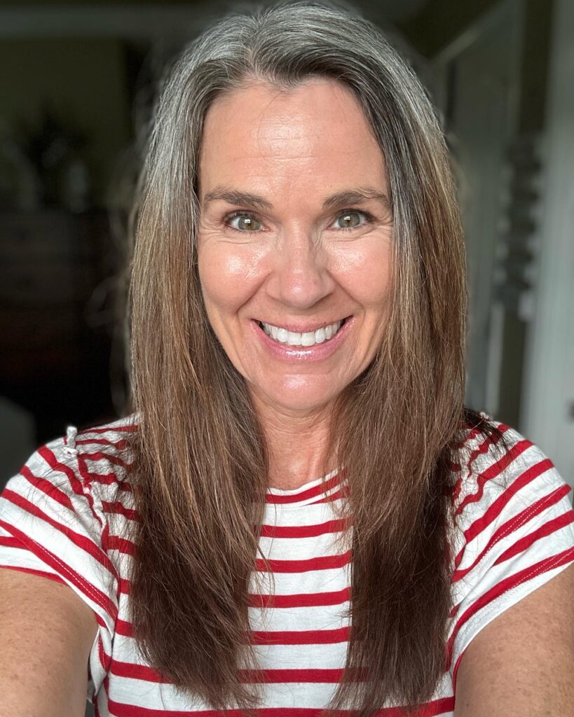A woman with long straight dark brown hair and natural silver roots wearing a red and white striped shirt.