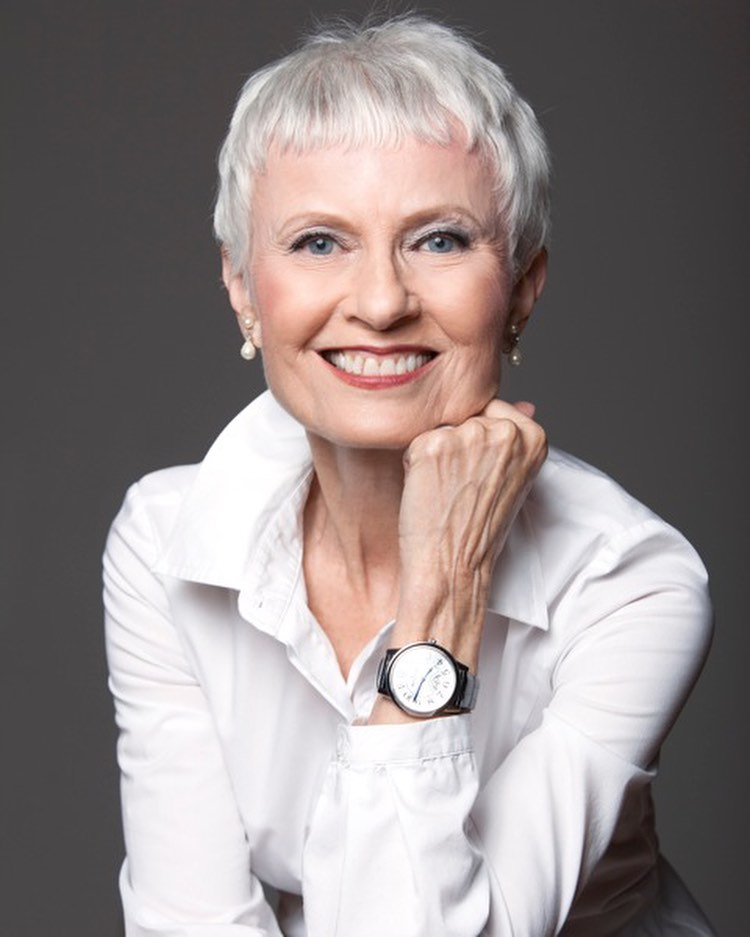 A woman with short silver hair in a textured pixie cut with soft bangs and a white collared shirt.