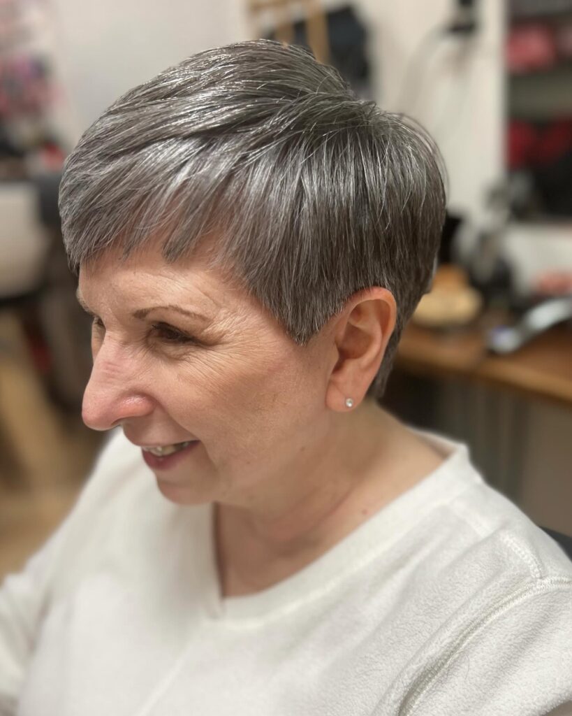 A woman with a short slate grey pixie haircut featuring soft micro bangs and a sleek tapered finish around the ears.