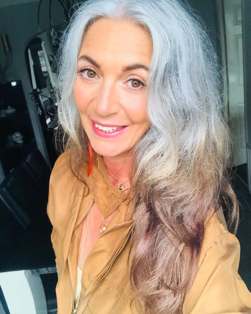 A woman with long wavy hair featuring a mix of natural silver roots and warm blonde ends.