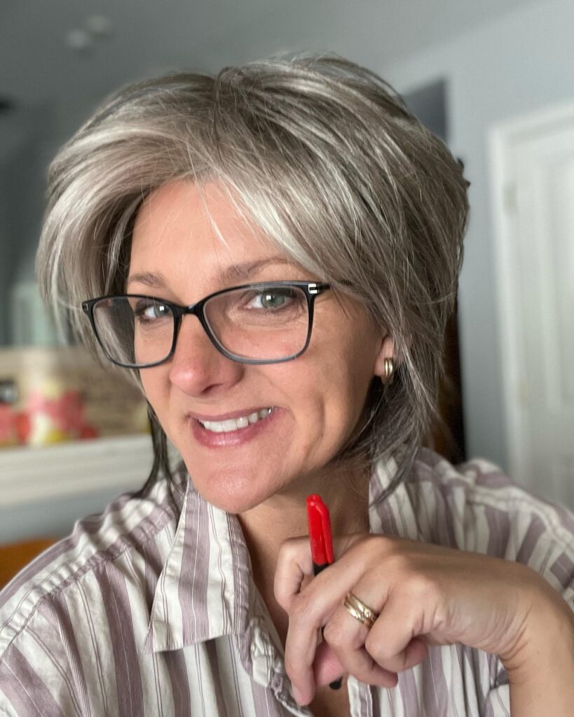 A woman wearing glasses with a voluminous ashy grey shaggy hairstyle featuring feathered layers and a soft fringe.