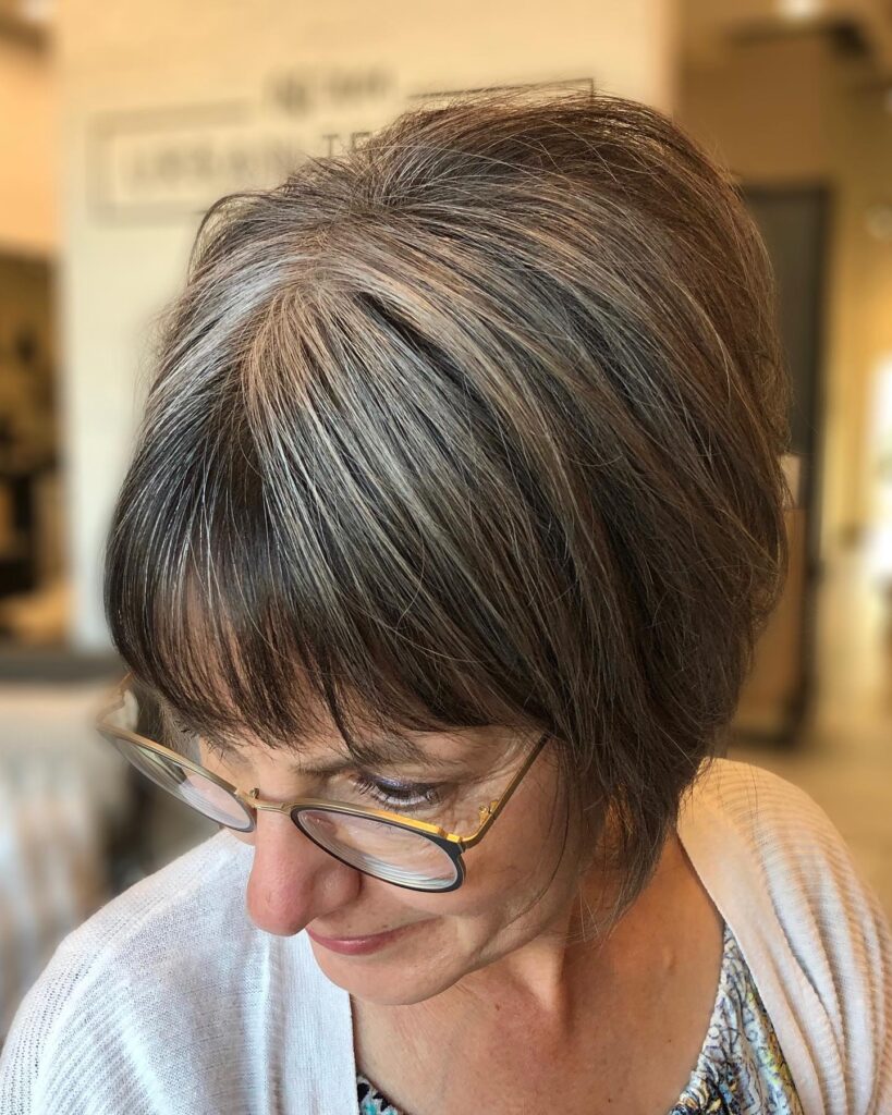 A woman with a chin length salt and pepper bob featuring thin wispy bangs and natural grey highlights.