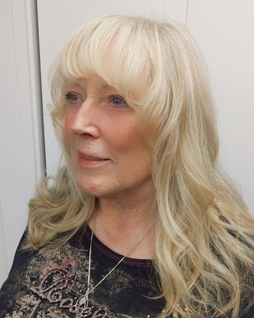A woman with long wavy blonde hair featuring thick rounded bangs and face framing layers.