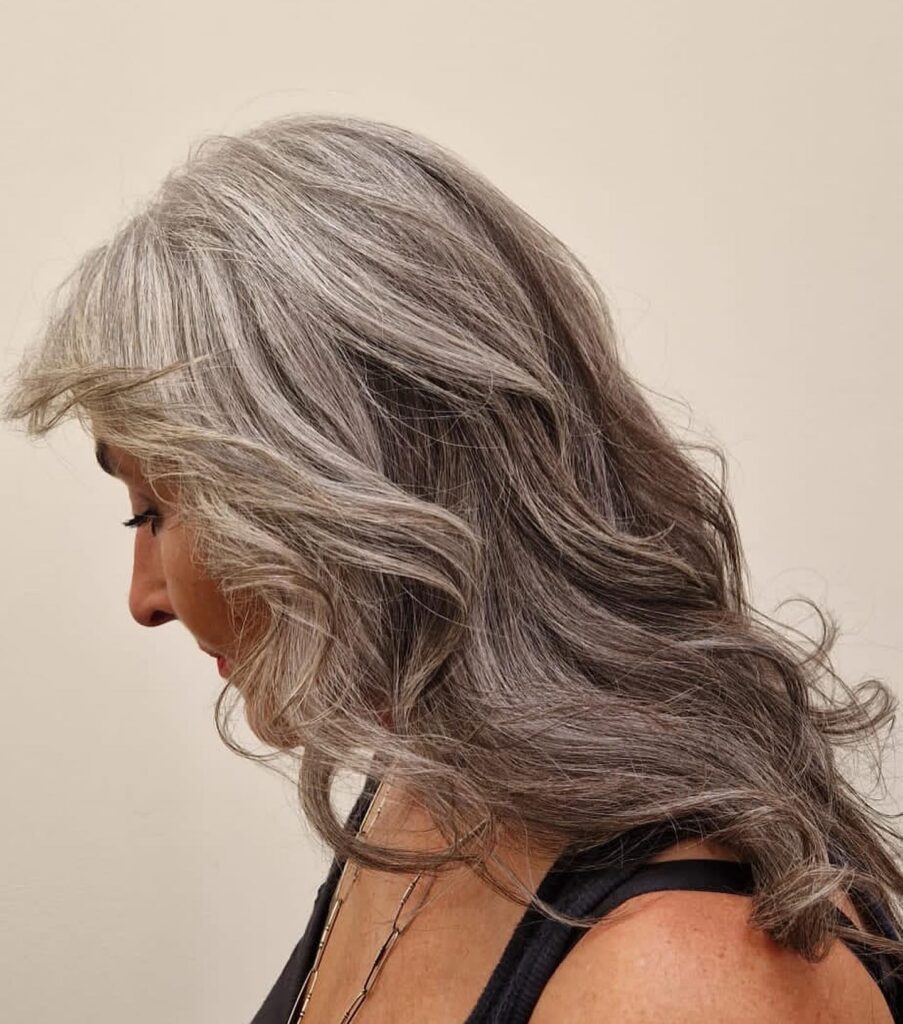 A woman with layered steel grey hair featuring bright silver highlights styled with large waves and a soft swept back fringe.