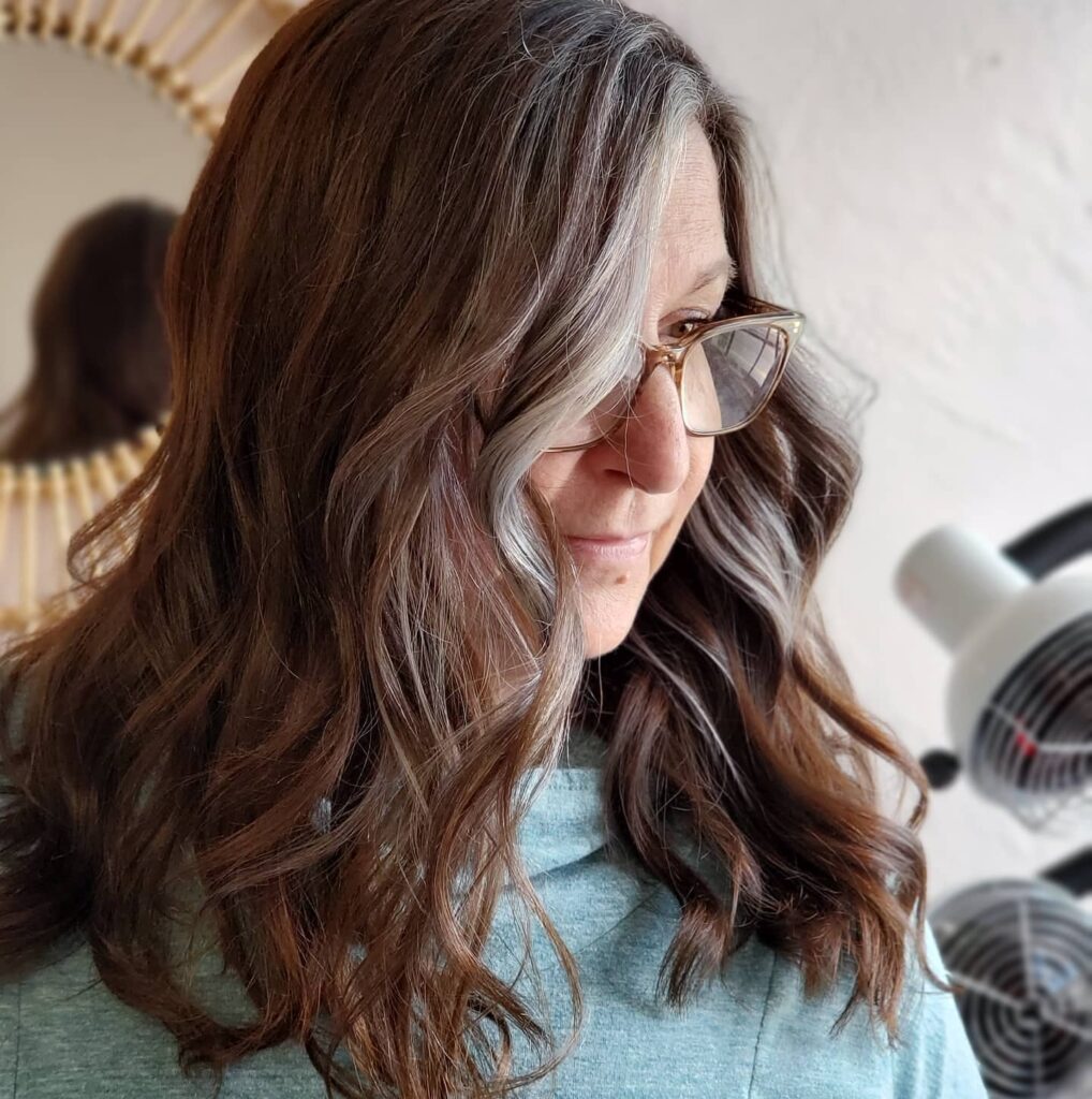 A woman with long wavy brunette hair featuring a prominent natural silver streak framing her face.