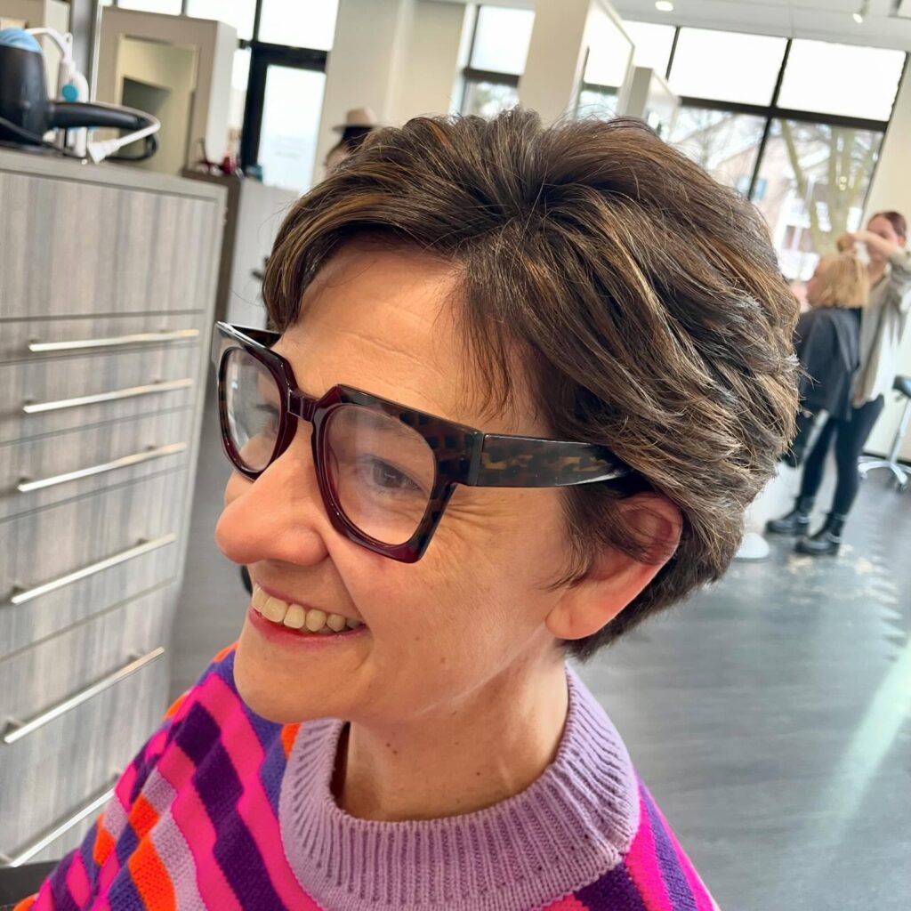 A woman wearing stylish glasses showcases a voluminous brunette pixie cut with warm highlights and textured layers.