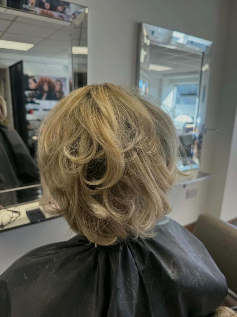 The back view of a woman with a voluminous short golden blonde hairstyle featuring soft bouncy curls and layered texture.