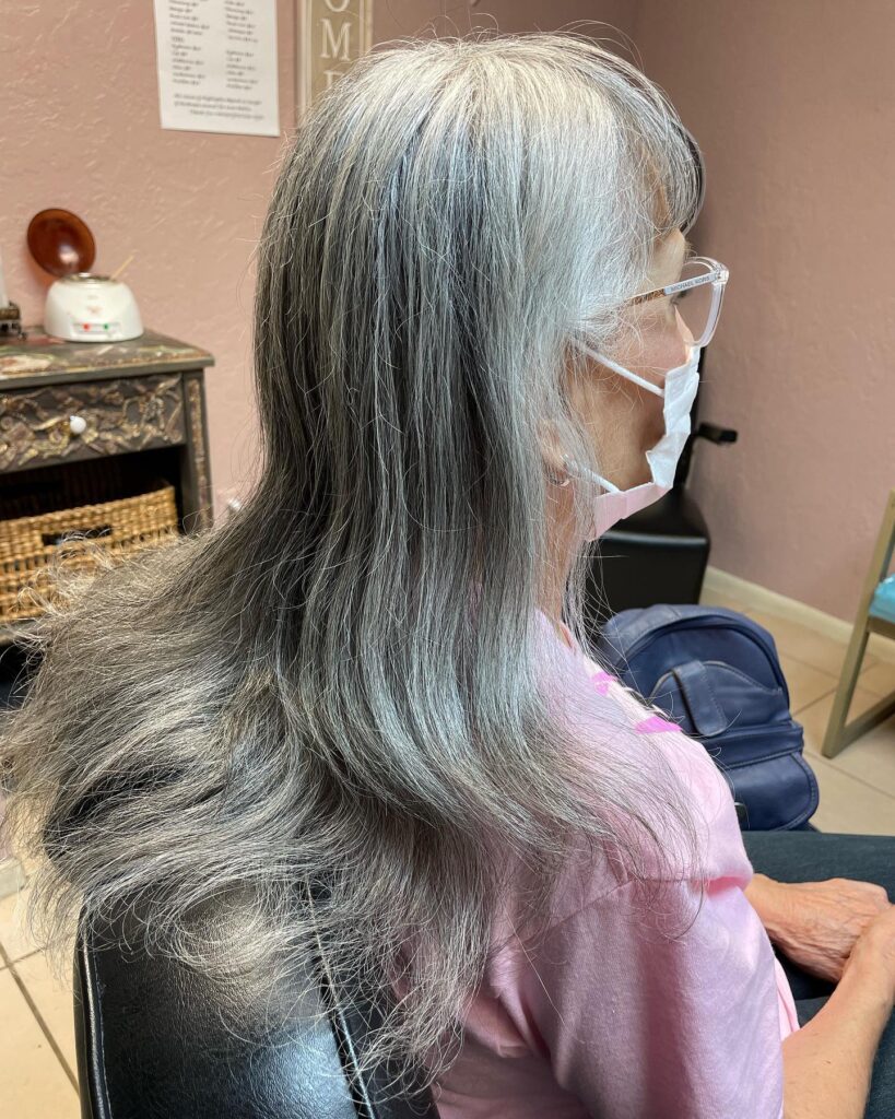 A woman with long thick salt and pepper silver hair featuring soft layers and wispy front bangs.